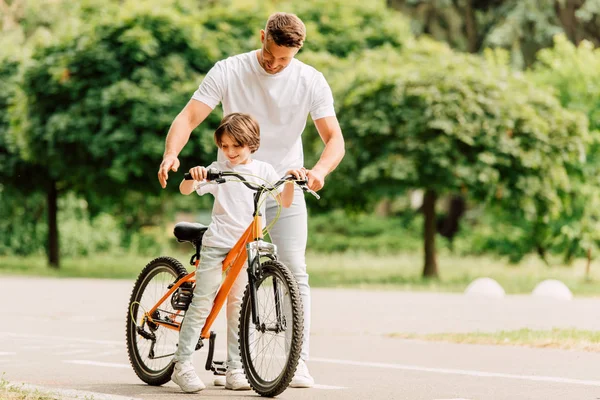 Visão de comprimento total do pai e do filho olhando na bicicleta enquanto de pé na estrada — Fotografia de Stock