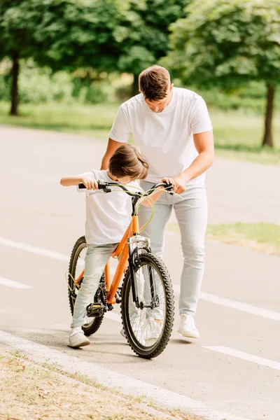 Visão de comprimento total do filho tentando sentar-se na bicicleta enquanto o pai segurando bicicleta — Fotografia de Stock