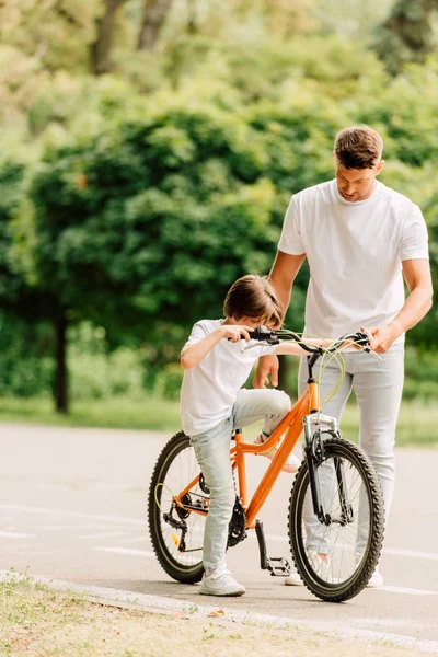 Vista integrale del figlio che cerca di sedersi in bicicletta mentre il padre tiene in mano la bici e guarda il bambino — Foto stock