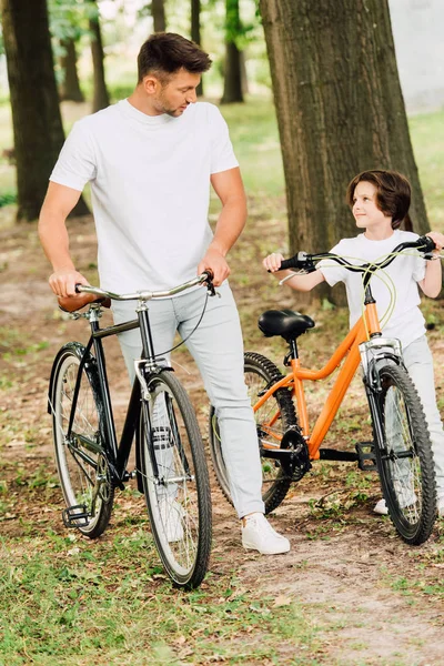 Vista completa de padre e hijo caminando en el parque con bicicletas y mirándose el uno al otro - foto de stock
