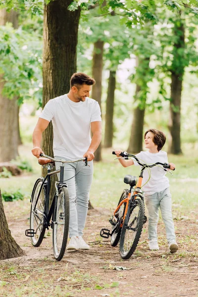 Vue pleine longueur de père et fils heureux se regardant et souriant tout en marchant avec des vélos dans la forêt — Photo de stock