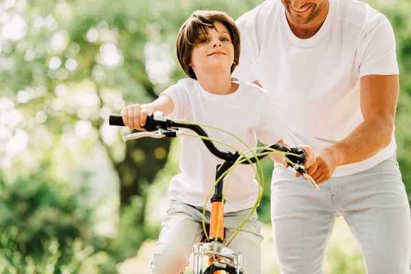 Visão cortada do pai ajudando o filho enquanto criança andando de bicicleta e olhando para a frente — Fotografia de Stock