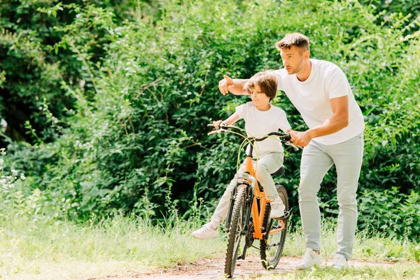 Pleine longueur vue de fils à vélo tandis que le père aidant enfant et pointant du doigt — Photo de stock