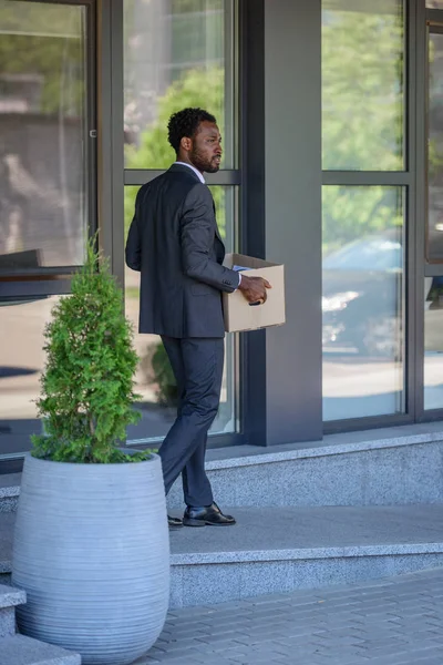 Serious african american businessman carrying carton box while walking near office building — Stock Photo