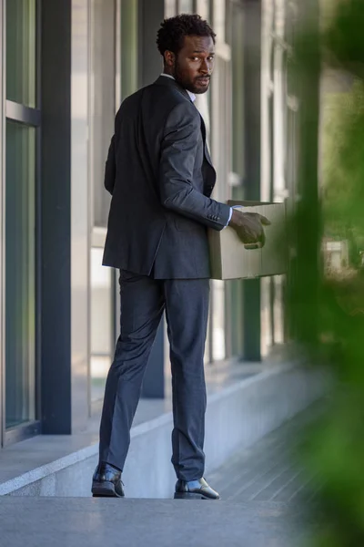 Enfoque selectivo del empresario afroamericano despedido mirando hacia atrás mientras sostiene la caja de cartón - foto de stock