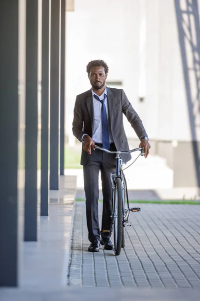 Thoughtful african american businessman walking near office building with bicycle — Stock Photo
