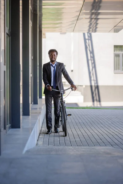 Cheerful african american businessman with bike walking near office building — Stock Photo