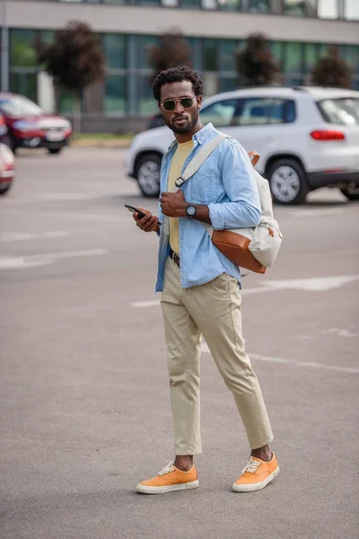 Bel homme afro-américain en lunettes de soleil tenant smartphone tout en se tenant debout sur le parking — Photo de stock