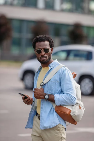 Hombre afroamericano sosteniendo teléfono inteligente y mirando a la cámara mientras está de pie en el aparcamiento - foto de stock