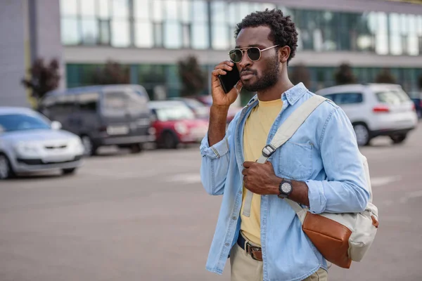 Bel homme afro-américain parlant sur smartphone tout en se tenant debout sur le parking et en regardant loin — Photo de stock