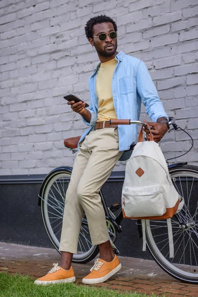 Elegante smartphone africano americano e guardando lontano mentre in piedi con la bicicletta vicino alla parete — Foto stock