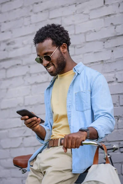 Cheerful african american man smiling while standing near brick wall and using smartphone — Stock Photo