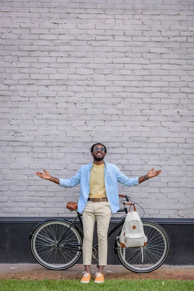 Feliz homem afro-americano gesticulando enquanto estava perto da parede de tijolo com bicicleta — Fotografia de Stock