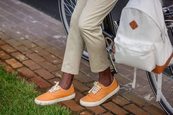 Vista recortada de hombre afroamericano con estilo de pie cerca de la bicicleta con mochila - foto de stock