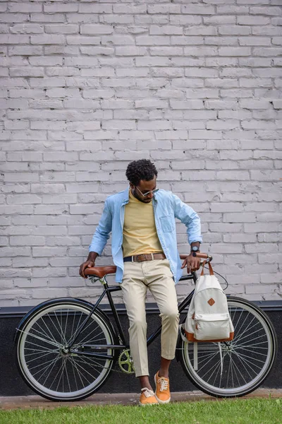 Guapo, elegante hombre afroamericano de pie con bicicleta cerca de la pared de ladrillo - foto de stock