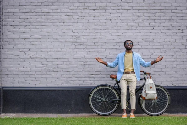 Excité homme afro-américain geste tout en se tenant près du mur de briques avec vélo — Photo de stock