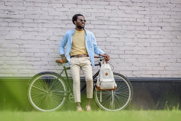 Hombre afroamericano elegante mirando hacia otro lado mientras que está parado con la bicicleta cerca de la pared de ladrillo - foto de stock