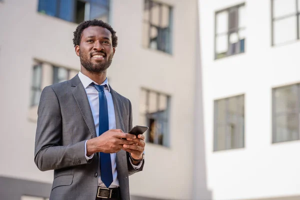 Alegre hombre de negocios afroamericano mirando hacia otro lado mientras sostiene el teléfono inteligente - foto de stock