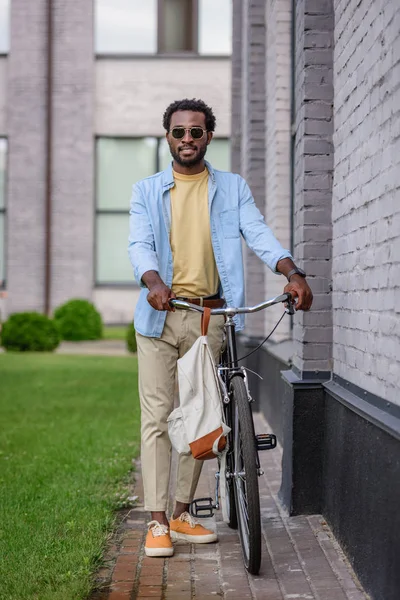 Красивий, стильний афроамериканський чоловік в сонцезахисних окулярах дивиться на камеру, стоячи з велосипедом біля цегляної стіни — стокове фото