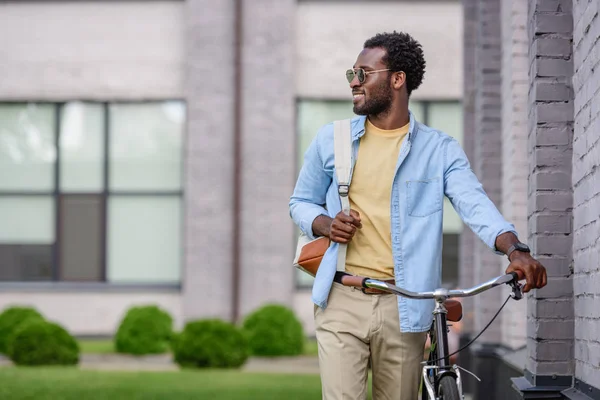 Handsome african american man looking away while standing with bike near brick building — Stock Photo