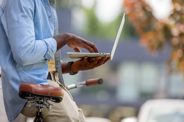 Vista recortada del hombre afroamericano usando el ordenador portátil mientras está de pie cerca de la bicicleta - foto de stock