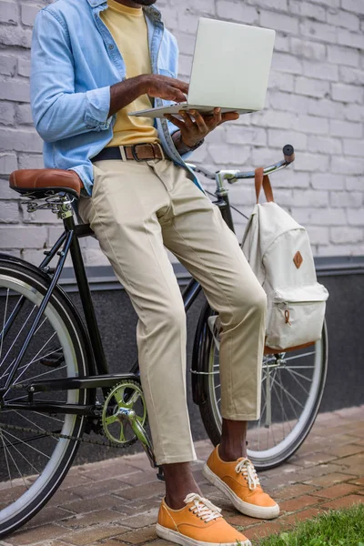 Vue partielle de l'homme afro-américain en utilisant un ordinateur portable tout en se tenant près du vélo — Photo de stock