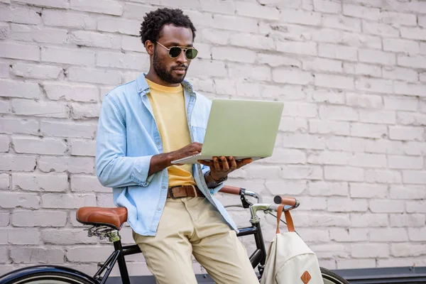 Attentif, élégant homme afro-américain en utilisant un ordinateur portable tout en se tenant près du mur de briques près de vélo — Photo de stock