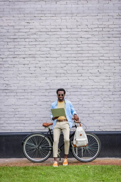 Stylish african american man standing by brick wall near bicycle and using smartphone — Stock Photo