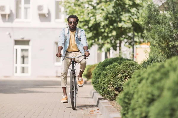 Handsome african american man riding bicycle along sunny street and green plants — Stock Photo