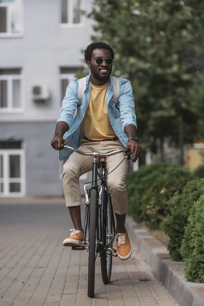Alegre, hombre afroamericano elegante sonriendo mientras que monta bicicleta - foto de stock