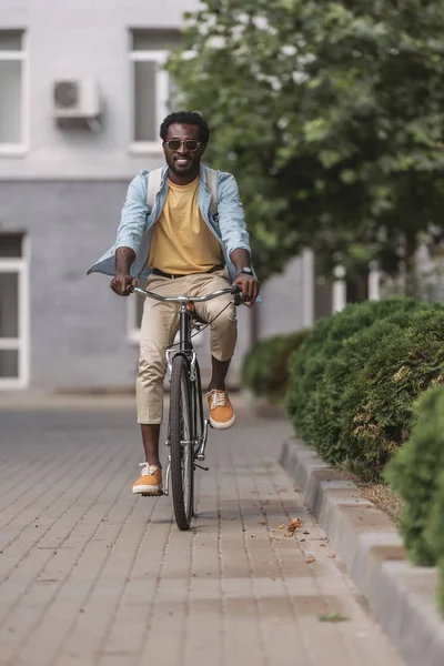 Guapo, elegante hombre afroamericano sonriendo mientras monta bicicleta a lo largo de la calle - foto de stock