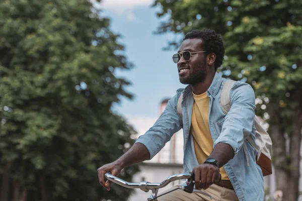 Bonito afro-americano homem em óculos de sol olhando para longe e sorrindo ao andar de bicicleta — Fotografia de Stock