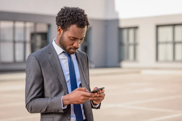 Atento hombre de negocios afroamericano en traje usando teléfono inteligente en la calle - foto de stock