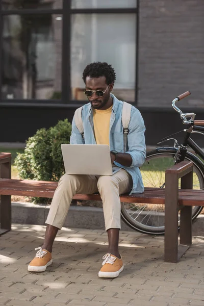 Élégant homme afro-américain en utilisant un ordinateur portable tout en étant assis sur un banc près de vélo — Photo de stock