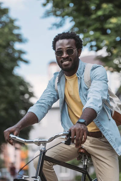 Homme afro-américain joyeux en lunettes de soleil souriant à la caméra tout en faisant du vélo — Photo de stock