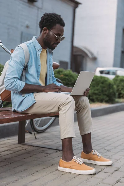 Stylish african american man in sunglasses using laptop while sitting on bench — Stock Photo