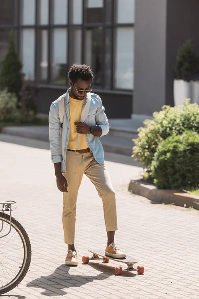 Stylish african american man in sunglasses standing near longboard on sunny street — Stock Photo