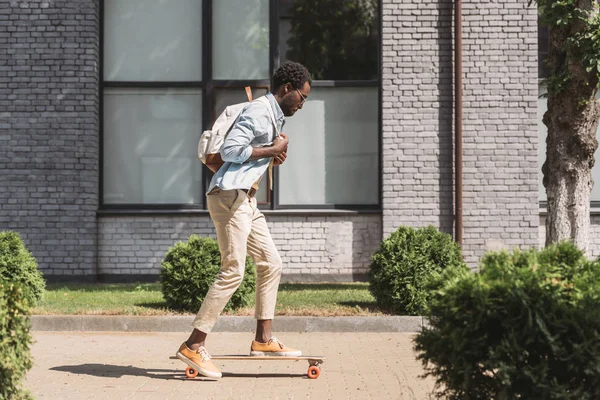 Homem americano africano elegante com mochila montando longboard na rua ensolarada — Fotografia de Stock