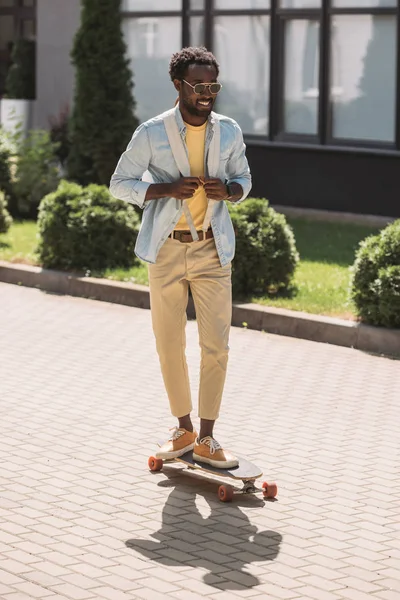 Joyeux, élégant homme afro-américain souriant tandis que longboard sur la rue ensoleillée — Photo de stock