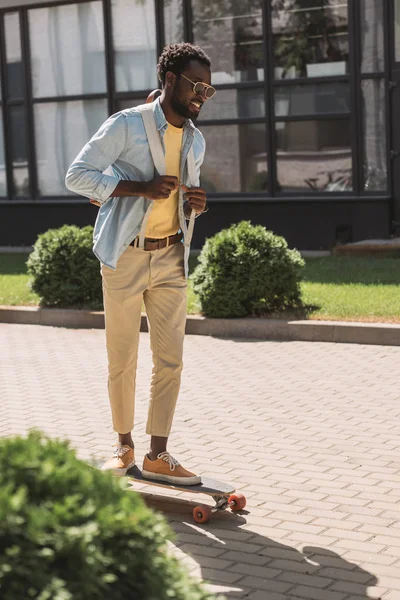Bonito, elegante homem afro-americano sorrindo enquanto montava longboard na rua ensolarada — Fotografia de Stock