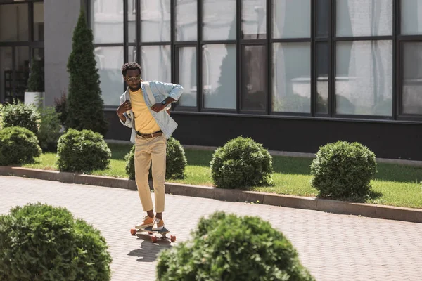 Стильный африканский американец, катающийся на длинной доске на солнечной улице возле зеленых кустов — стоковое фото
