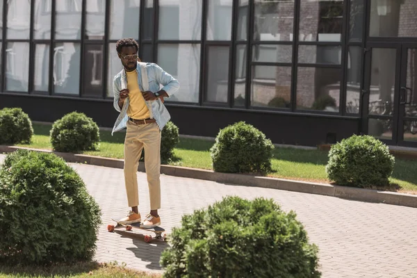Handsome african american man longboarding on sunny street near building and green bushes — Stock Photo