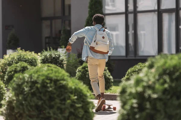 Vista trasera del hombre afroamericano con mochila montando longboard cerca de arbustos verdes en la calle - foto de stock