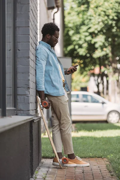 Homem americano africano elegante usando smartphone enquanto está perto do edifício com longboard — Fotografia de Stock