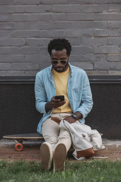 Stylish african american man using smartphone while sitting on longboard near wall — Stock Photo