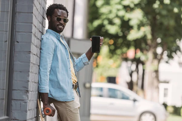 Guapo afroamericano hombre en gafas de sol mostrando teléfono inteligente con pantalla en blanco y mirando a la cámara - foto de stock