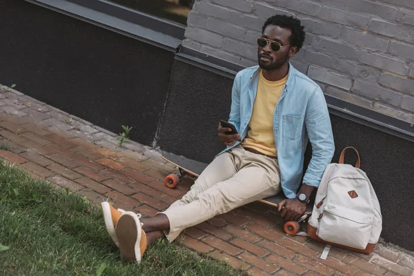 Bonito, elegante homem americano africano sentado no longboard e segurando smartphone — Fotografia de Stock