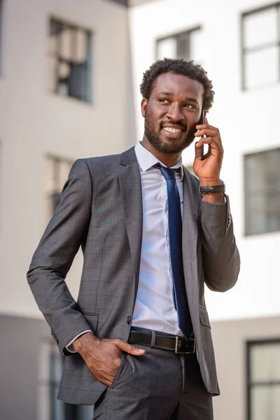 Alegre afroamericano hombre de la mano en el bolsillo y mirando hacia otro lado mientras habla en el teléfono inteligente - foto de stock