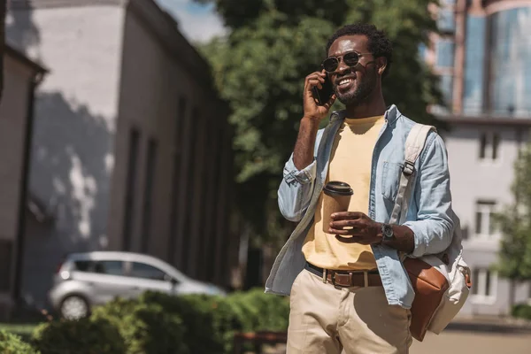 Stylischer, fröhlicher afrikanisch-amerikanischer Mann, der mit dem Smartphone spricht und wegschaut, während er Kaffee to go in der Hand hält — Stockfoto