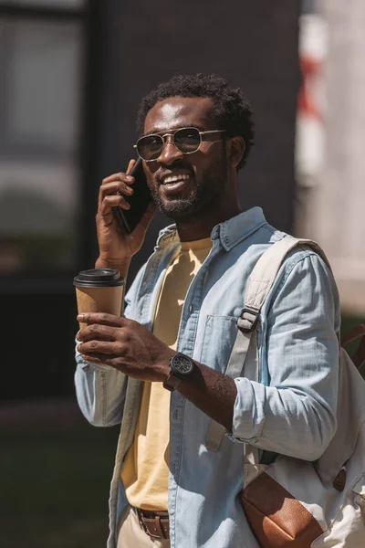 Sonriente afroamericano hombre hablando en el teléfono inteligente mientras sostiene el café para ir y mirar hacia otro lado - foto de stock